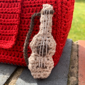 Pu&ograve; includere: Una borsa rossa all'uncinetto con una piccola chitarra all'uncinetto attaccata al lato. La chitarra &egrave; beige con corde marroni.