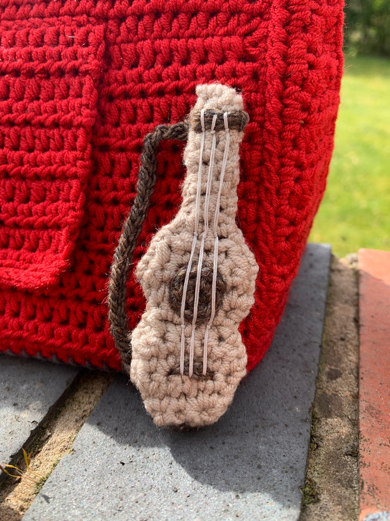Pu&ograve; includere: Una borsa rossa all'uncinetto con una piccola chitarra all'uncinetto attaccata al lato. La chitarra &egrave; beige con corde marroni.