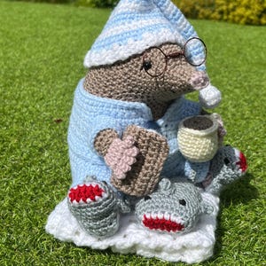 May include: A crocheted mole wearing a blue and white striped sleep cap, blue pajamas, and round glasses, holding a cup of coffee and sitting on a white blanket with two crocheted shark toys.