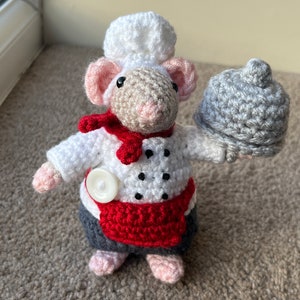 May include: A crocheted mouse wearing a chef's hat and apron, holding a crocheted cloche. The mouse is white with black polka dots and has a red scarf.