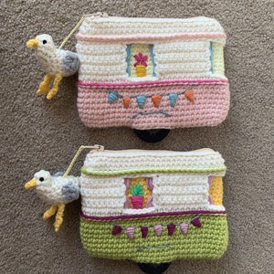 May include: Two crocheted caravan coin purses with a seagull charm. The top purse is pink with white trim and the bottom purse is green with white trim. Both purses have a window with a potted plant and a string of bunting.
