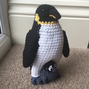 May include: A crocheted penguin toy with black and white fur, a yellow beak, and a smaller crocheted penguin chick underneath.