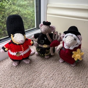 May include: Three crocheted mouse figurines. The left mouse wears a red tunic and tall black hat. The centre mouse wears a black jacket and green plaid kilt with a small bagpipe. The right mouse wears a black hat and red vest with a yellow flower.