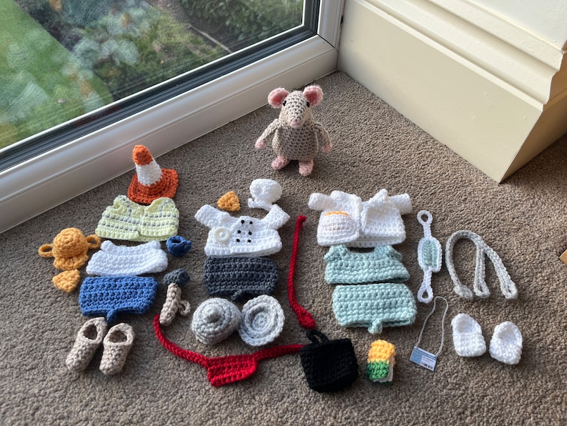 Op de afbeelding: Een verzameling gehaakte outfits en accessoires voor een klein knuffeldier. De outfits omvatten een koksuniform, een doktersjas, een bouwvakkersvest en een badpak. De accessoires omvatten een hoed, een stethoscoop, een gereedschapsgordel en een tas.