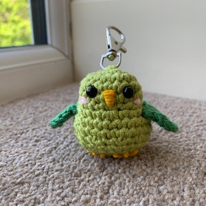 May include: A green crocheted parakeet keychain with yellow beak and black eyes. The parakeet has pink cheeks and green wings.