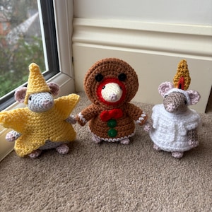 Op de afbeelding: Drie handgemaakte gehaakte muizen in feestelijke outfits. Eén muis draagt een geel sterrenkostuum en hoed, een andere is verkleed als peperkoekenmannetje met een rode strik, en de derde heeft een witte jurk en een hoorn.