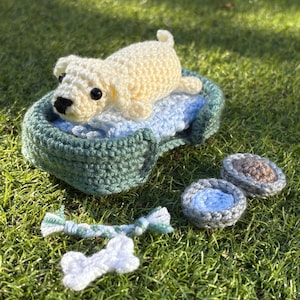 May include: A cream-colored crocheted puppy figurine resting in a dark green crocheted bed with a light blue cushion. Included are two small gray crocheted bowls, a white crocheted bone, and a braided white and green rope toy accessory.