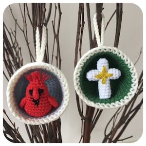 May include: Two crocheted ornaments hanging on a branch. One ornament is white with a red cardinal inside. The other ornament is green with a white cross inside.