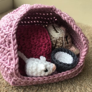 May include: A pink crocheted sleeping bag with a white crocheted mouse, a brown crocheted cat, and a gray crocheted bowl inside.