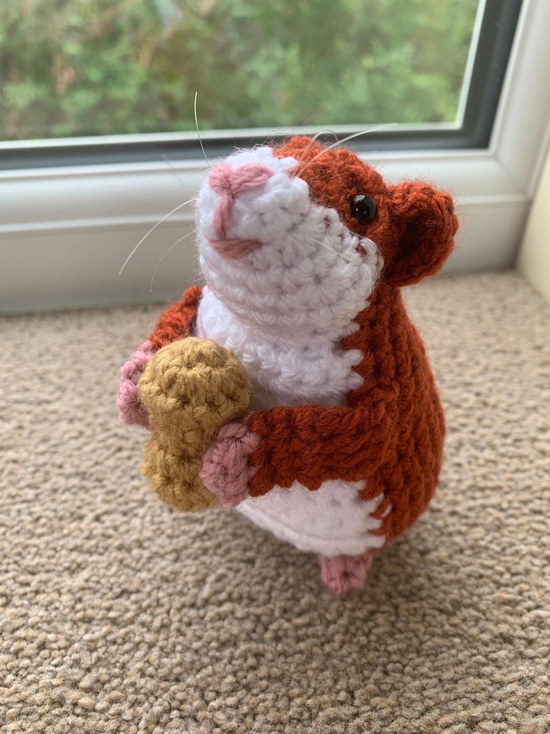 May include: A crocheted hamster toy holding a peanut in its paws. The hamster is brown, white, and pink.