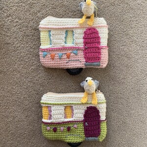 May include: Two crocheted campervan coin purses with a crocheted seagull on top. The top campervan is pink with yellow windows and a pink door. The bottom campervan is green with yellow windows and a purple door.
