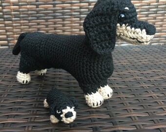 May include: Two crocheted dachshund dogs, one black and tan with white paws, the other smaller and all black, on a brown wicker surface.