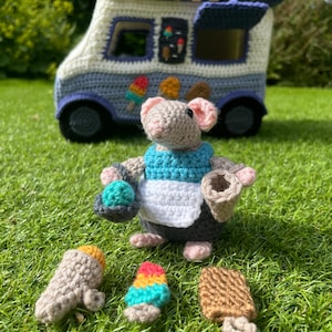 May include: A crocheted mouse wearing a blue and white apron holds a crocheted ice cream cone. The mouse is standing in front of a crocheted ice cream van. There are three crocheted ice cream cones on the ground.