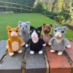 May include: Five crocheted cat toys in different colours, including orange, grey, black, brown, and grey with white accents. The toys are arranged in a row on a brick surface.