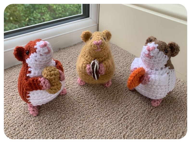 May include: Three crocheted hamster plushies holding different colored crocheted treats. The hamsters are brown, orange, and white with pink noses and feet.