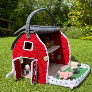 Könnte beinhalten: Ein roter Häkel-Stall mit einem grauen Dach und einem weißen Lattenzaun. Der Stall hat eine Tür, die sich öffnen lässt, und ein Fenster. Im Stall befinden sich kleine Häkeltiere, darunter eine Kuh, ein Schwein und Hühner.
