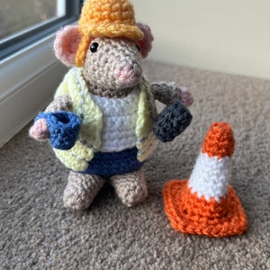 Op de afbeelding: Een gehaakte muis met een gele helm, een gele vest, een blauwe spijkerbroek en een blauw en zwart gereedschap in de hand. De muis staat naast een oranje-witte gehaakte verkeerskegel.