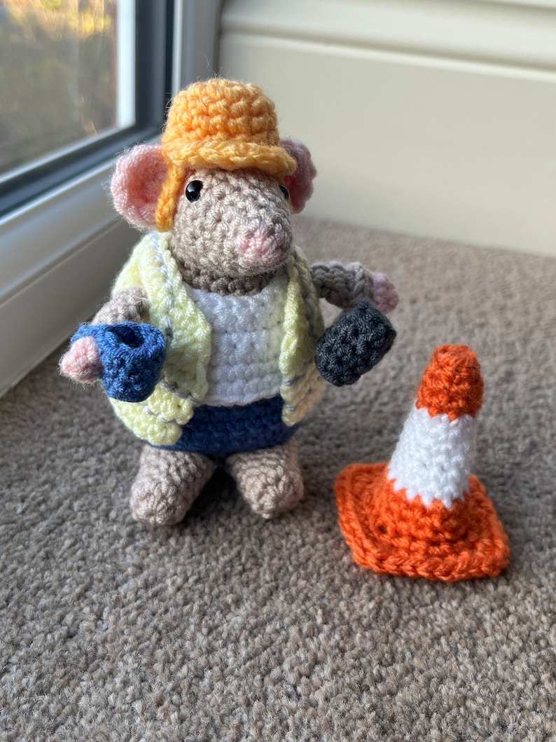 Op de afbeelding: Een gehaakte muis met een gele helm, een gele vest, een blauwe spijkerbroek en een blauw en zwart gereedschap in de hand. De muis staat naast een oranje-witte gehaakte verkeerskegel.