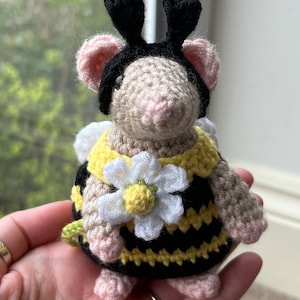 May include: A crocheted bee-themed plush toy with black antennae, yellow and black striped body, and a white flower. The toy is sitting in a person's hand.