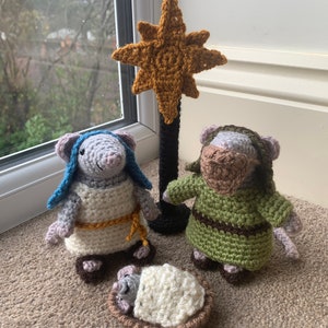 Peut inclure: Une scène de la nativité en crochet avec une souris grise, une souris verte et un bébé souris blanc dans une crèche brune. Une étoile dorée est placée sur un support noir derrière les figurines.