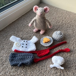 Op de afbeelding: Een gehaakte grijze muis met roze oren en een witte knoop op zijn borst. De muis is omringd door gehaakte kledingstukken, waaronder een wit shirt, een grijze broek, een rood vest, een witte muts en een grijs bord met een geel gehaakt stuk voedsel.