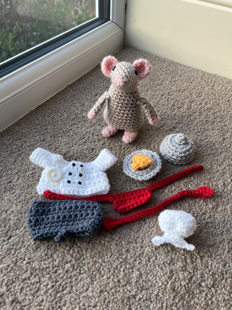 Op de afbeelding: Een gehaakte grijze muis met roze oren en een witte knoop op zijn borst. De muis is omringd door gehaakte kledingstukken, waaronder een wit shirt, een grijze broek, een rood vest, een witte muts en een grijs bord met een geel gehaakt stuk voedsel.