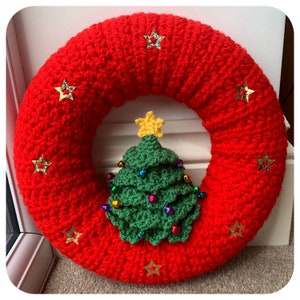 May include: A red crocheted Christmas wreath with a green crocheted Christmas tree in the centre. The wreath is decorated with gold glitter stars.