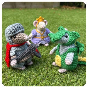 Può includere: Tre figure amigurumi all'uncinetto: un topo cavaliere in armatura grigia con sciarpa e scudo rossi, un topo con un vestito viola e corona d'oro e un drago verde. Tutti su uno sfondo di erba verde.
