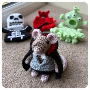 May include: A selection of crocheted toys: a skeleton, a devil, an alien, a caterpillar, and a mouse in a cape and vest. The toys are crafted from various coloured yarns and are arranged on a neutral carpet, showcasing a range of textures and designs.