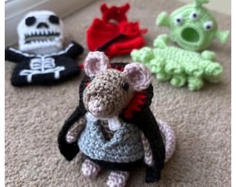 May include: A collection of handmade crochet toys, including a skeleton, a devil, an alien, a caterpillar, and a mouse dressed in a cape and vest. The toys are made with various colors of yarn and are displayed on a neutral-colored carpet.