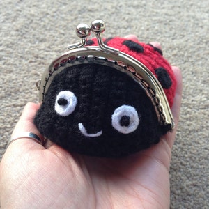 May include: A black and red crocheted ladybug coin purse with a silver clasp and white eyes. The purse has a smiling mouth.