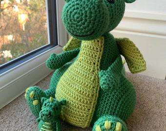 May include: Two crocheted green dragons, one large and one small, sit on a neutral-colored carpet. The larger dragon has a darker green body with lighter green accents on its belly, wings, and feet. The smaller dragon is a miniature version.