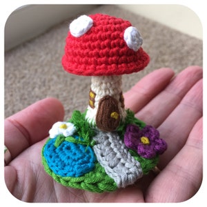 May include: A crocheted mushroom house with a red roof, white spots, and a brown door. The house is surrounded by green grass, a blue pond, a gray path, and purple flowers.