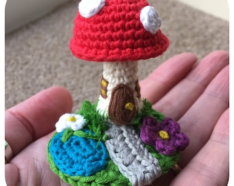 May include: A crocheted mushroom house with a red roof, white spots, and a brown door. The house is surrounded by green grass, a blue pond, a gray path, and purple flowers.