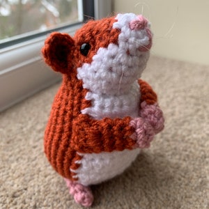 May include: A crocheted hamster toy with brown, white, and pink fur. The hamster is standing on its hind legs with its front paws raised.