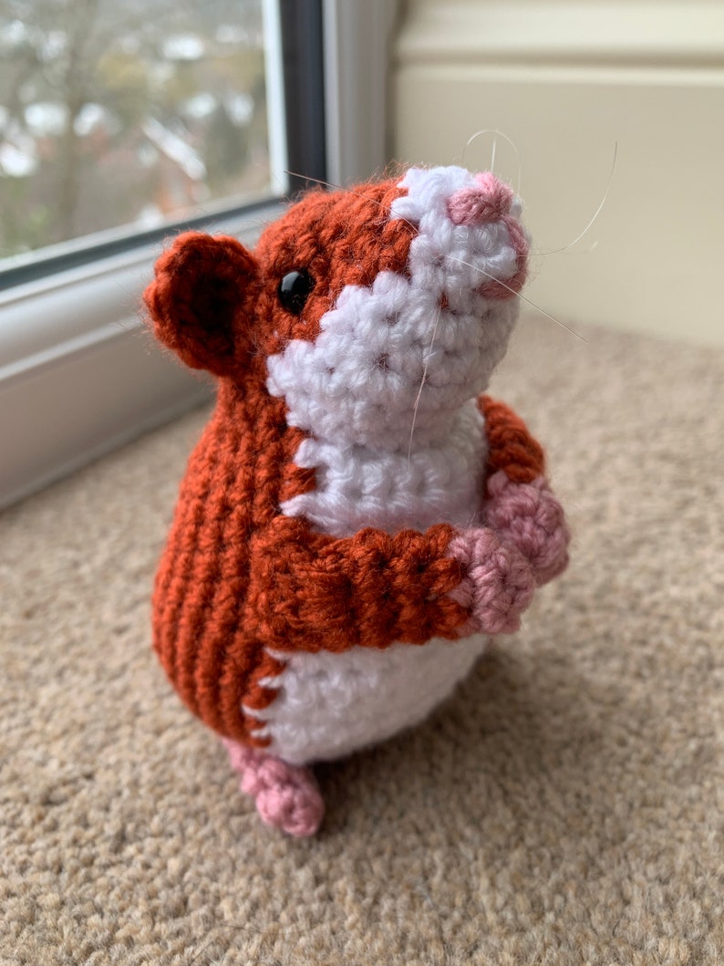 May include: A crocheted hamster toy with brown, white, and pink fur. The hamster is standing on its hind legs with its front paws raised.
