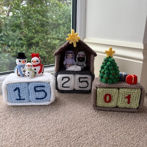 May include: Three handmade crochet perpetual calendars. The left one is white with blue blocks showing 15 and three snowmen. The center one is brown with dark gray blocks showing 25 and a nativity scene. The right one is brown with green blocks showing 01, a Christmas tree, and two small gifts.