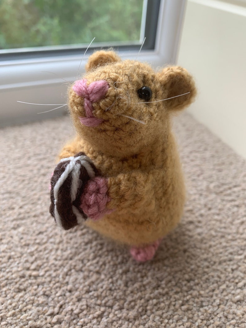 May include: A crocheted brown hamster holding a chocolate and white striped cookie. The hamster has pink ears, nose, and feet.