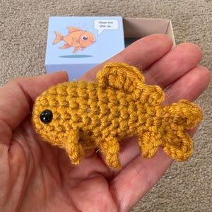 May include: A handmade, golden yellow crochet goldfish with a black eye is held in a hand. The fish has detailed fins and tail. A small box with a cartoon goldfish and the text "Please look after me..." is in the background.