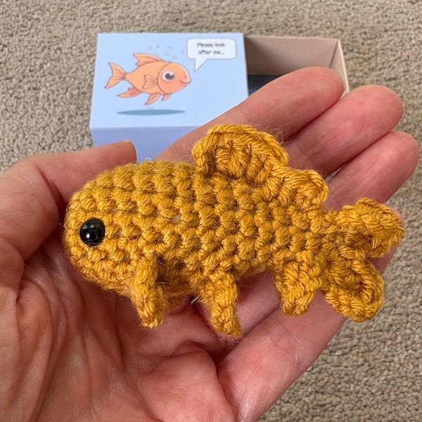 May include: A handmade, golden yellow crochet goldfish with a black eye is held in a hand. The fish has detailed fins and tail. A small box with a cartoon goldfish and the text "Please look after me..." is in the background.