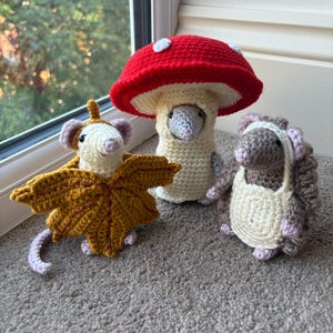 Op de afbeelding: Drie gehaakte figuren: een muis in een geel esdoornbladkostuum, een muis in een rood-witte paddenstoelhoed en een bruine muis met een crèmekleurig schort. Elk figuur heeft kleine oortjes, een spitse snuit en een gedetailleerde textuur van garen.