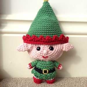 May include: A crocheted elf doll with a green hat, red trim, and a pink face. The elf is wearing a green and black outfit with a white belt.