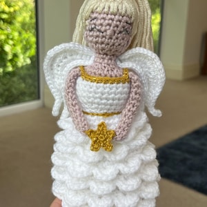 Op de afbeelding: Een gehaakte engelpop gemaakt van wit en roze garen, een gouden halo en een gouden ster. De engel draagt een witte jurk met een gouden rand en heeft blond garenhaar.