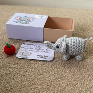 Op de afbeelding: Een gehaakte grijze olifant speelgoed naast een kleine rode gehaakte appel en een doos met een olifanten illustratie. Een kaart leest: "Mijn naam is: Edith. Ik hou van rolschaatsen & karamelijs. Kijk alsjeblieft naar me."