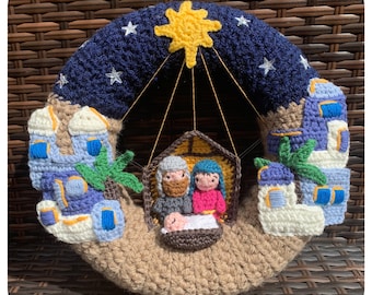 Puede incluir: Una corona de Navidad de ganchillo con una escena de la natividad con una estrella, un cielo azul noche y edificios blancos. La escena incluye un establo de madera marrón, una estrella amarilla y tres figuras: María, José y el niño Jesús.