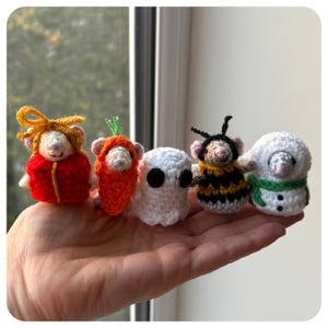 May include: Five miniature crochet figures including a mouse in a red gift box, a mouse in a carrot costume, a white ghost, a mouse in a bee costume, and a snowman with a green scarf. Each small stuffed character is crafted from yarn.