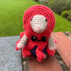 May include: A crocheted amigurumi toy in the shape of a small animal wearing a red hooded cloak. The toy has a pink nose, black eyes, and a small, crocheted tail.