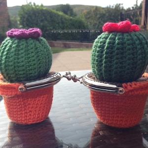 May include: Two crocheted cactus coin purses with a green cactus and a pink flower on top, each in an orange crocheted pot with a silver coin purse clasp.