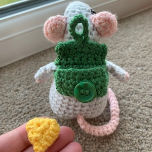 Pu&ograve; includere: Un topo bianco all'uncinetto con orecchie rosa e uno zaino verde all'uncinetto con un bottone verde. Il topo ha una coda rosa all'uncinetto. Un pezzo di formaggio giallo all'uncinetto &egrave; in primo piano.