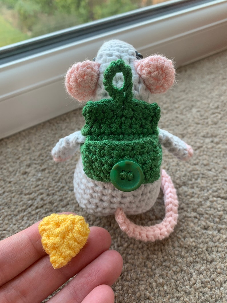 Pu&ograve; includere: Un topo bianco all'uncinetto con orecchie rosa e uno zaino verde all'uncinetto con un bottone verde. Il topo ha una coda rosa all'uncinetto. Un pezzo di formaggio giallo all'uncinetto &egrave; in primo piano.